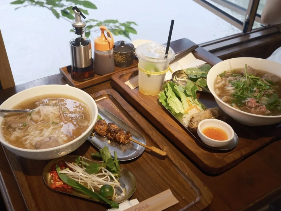 Bowls of Vietnamese noodle soup, sides and a drink by the window at PHO U.